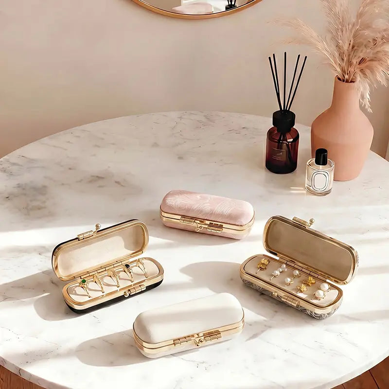 Four small vintage-style travel jewelry cases in pink embroidery, jacquard, black, and cream white leather arranged on a marble table with rings and earrings displayed.