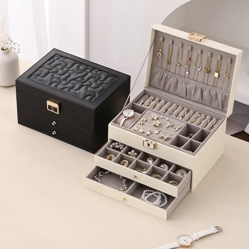 A closed black 3-tier jewelry box placed next to a fully open cream-white jewelry box with a grey velvet interior, displaying organized gold necklaces, rings, and earrings.