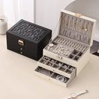 A closed black 3-tier jewelry box placed next to a fully open cream-white jewelry box with a grey velvet interior, displaying organized gold necklaces, rings, and earrings.