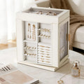 A stylish cream jewelry cabinet with a swivel base on a marble table next to a perfume bottle and a magazine, displaying organized earrings and rings in a cozy home environment.