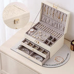 An open cream white 3-tier jewelry organizer box with a grey velvet lining, displaying gold necklaces, a white watch, floral earrings, and pearl bracelets, with a circular inset of the closed box.
