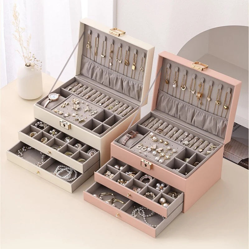 Two 3-tier jewelry organizer boxes in cream white and pink placed side-by-side on a table, both fully opened and showcasing organized watches, necklaces, earrings, and rings.
