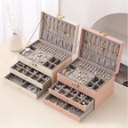 Two 3-tier jewelry organizer boxes in cream white and pink placed side-by-side on a table, both fully opened and showcasing organized watches, necklaces, earrings, and rings.