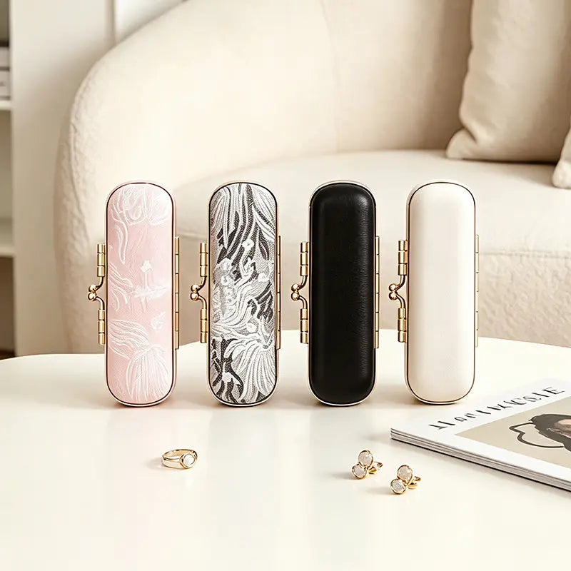 Four mini travel jewelry boxes standing vertically in a row showing variations of pink floral embroidery, grey jacquard fabric, sleek black leather, and smooth white leather with gold kiss-lock clasps.
