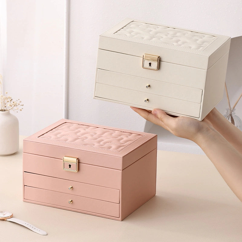 A person's hands holding up a cream-white 3-tier jewelry box, with a matching pink leather jewelry box sitting closed on the beige table next to it.
