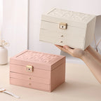 A person's hands holding up a cream-white 3-tier jewelry box, with a matching pink leather jewelry box sitting closed on the beige table next to it.