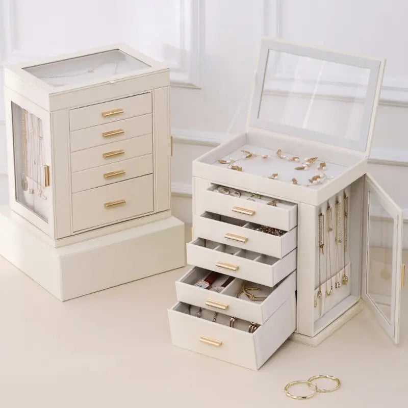 Two cream-white leather jewelry boxes with gold handles; one is open to show a top display compartment, five pull-out drawers, and a side glass door with hanging necklace hooks, displaying full storage capacity.