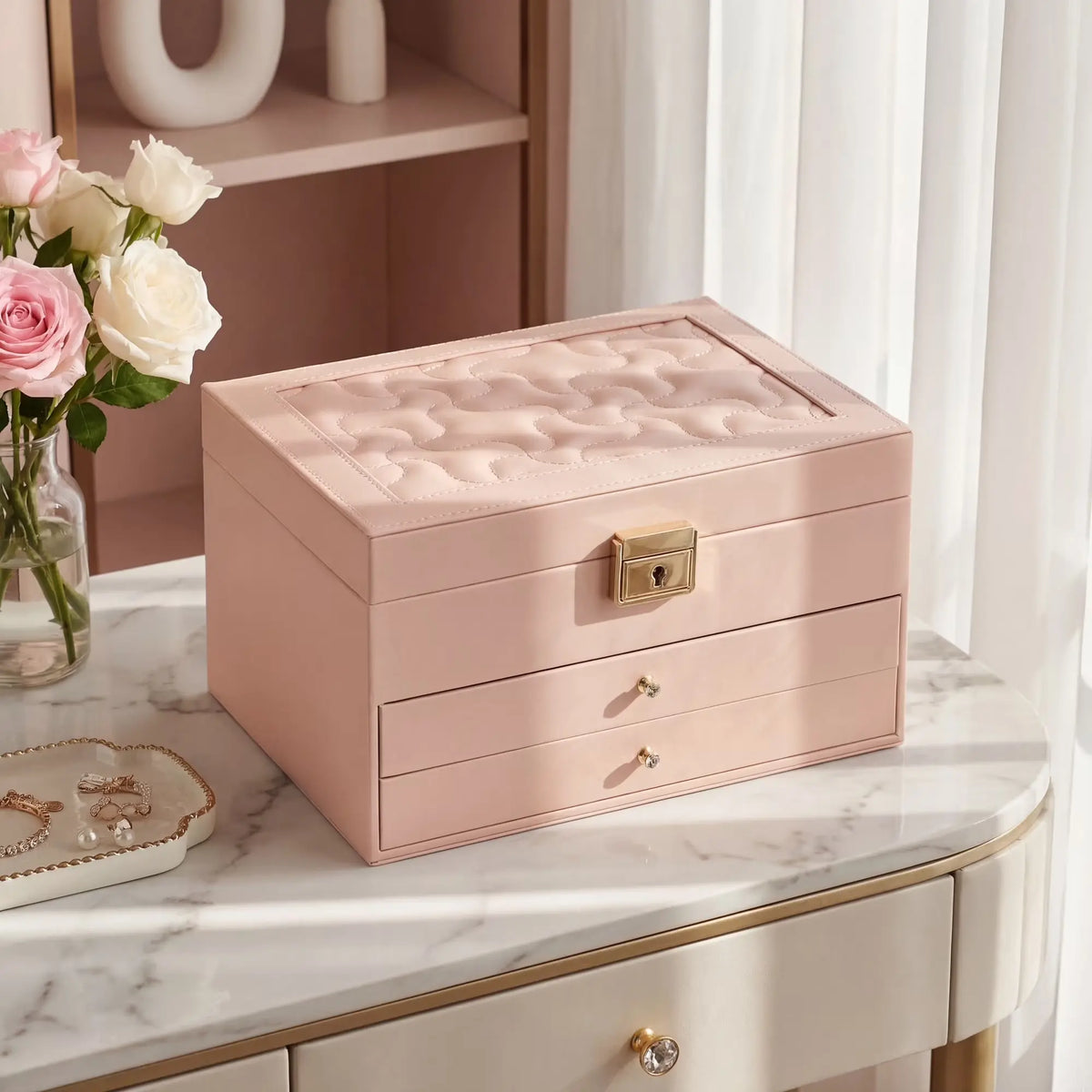 A closed pink leather 3-tier jewelry box sitting on a white marble vanity table, featuring a quilted embroidery top lid, gold lock clasp, and a vase of pink and white roses next to it.