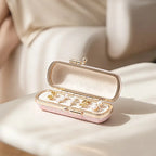 An open pink embroidered mini jewelry box displayed on a white chair, filled with several pairs of gold and pearl stud earrings.